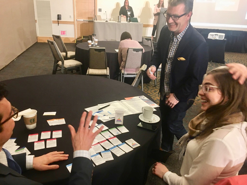 Two men and a woman discussing personalization using a card deck. They are seated at a round table in a hotel conference room. The workshop leaders, two women, are at a podium in the background.