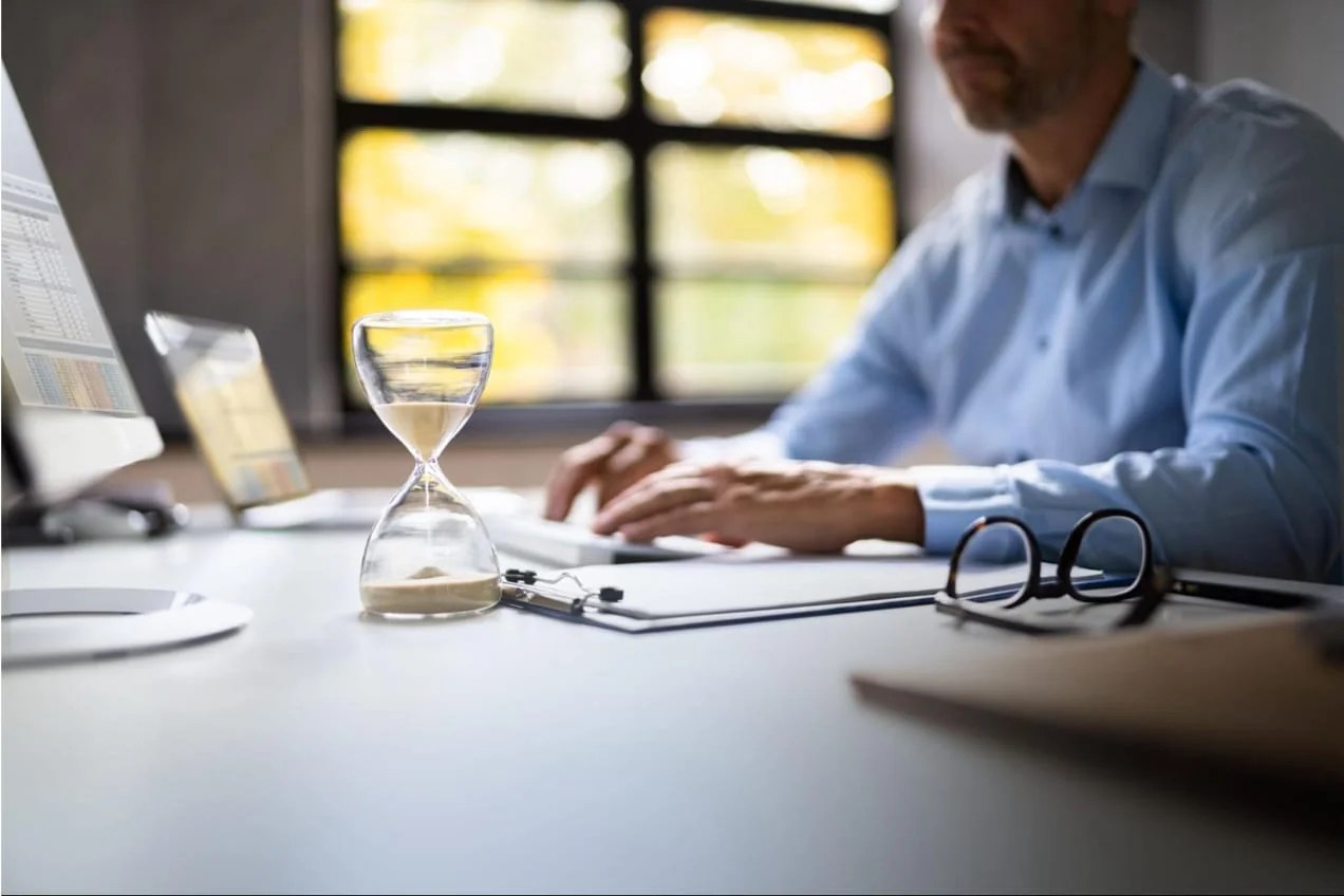 Man working on spreadsheets in an office with an hourglass halfway complete.