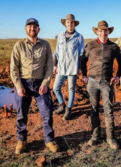 Three people, two men standing on either side of one woman, in a rural field wearing outdoor gear with work boots and wide brimmed hats.